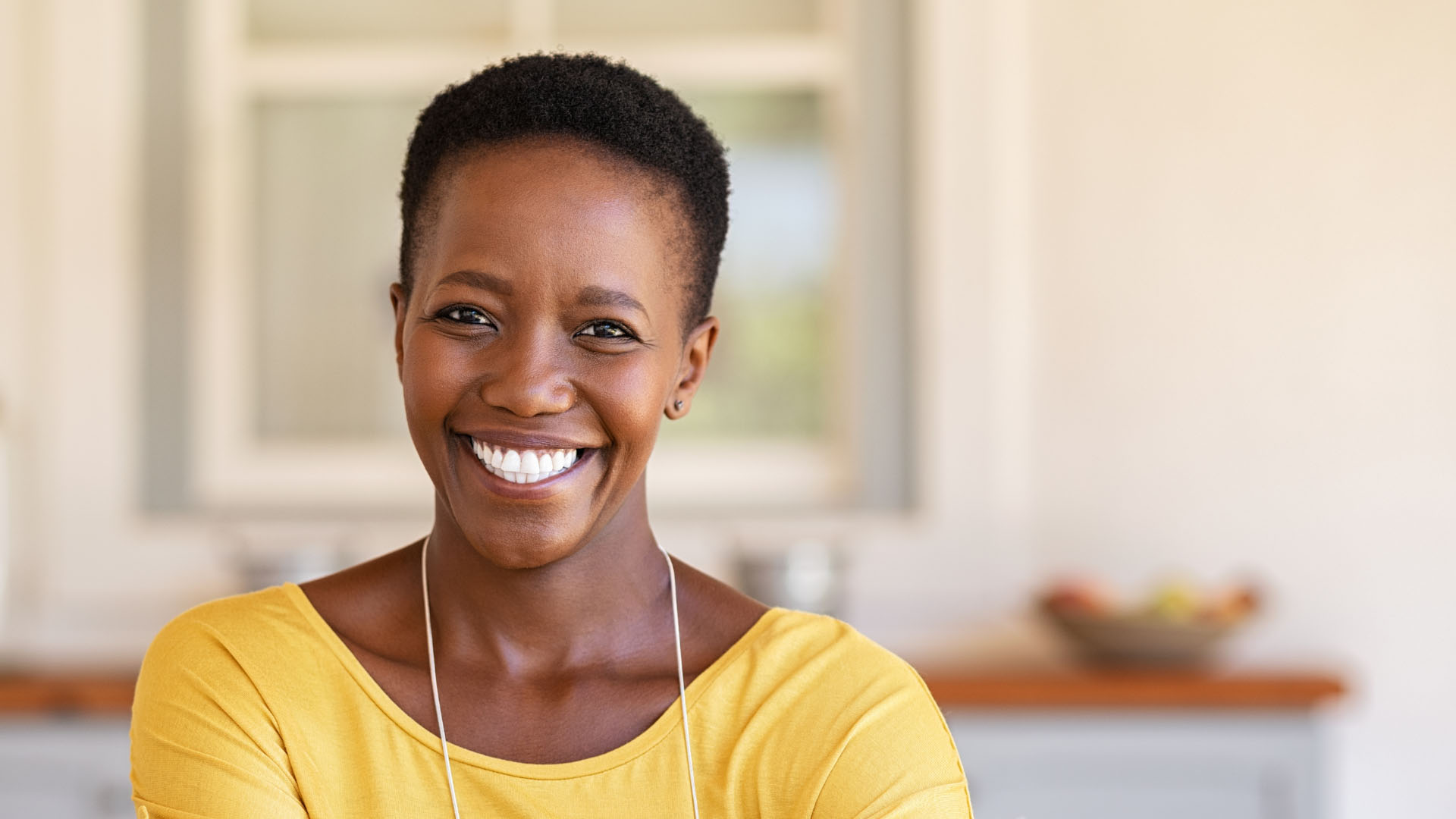 A smiling woman wearing a yellow top and earphones, photographed indoors in a bright kitchen setting.